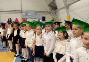 Uczniowie klas pierwszych w strojach galowych i biretach na głowie.