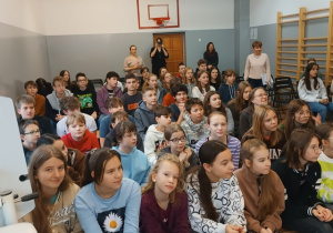 Uczniowie siedzący na ławeczkach w sali gimnastycznej podczas spotkania z aktorem.