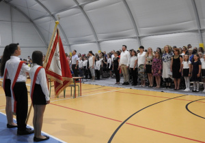 Uczniowie klas młodszych na zakończeniu roku szkolnego w hali sportowej, na pierwszym planie poczet sztandarowy.